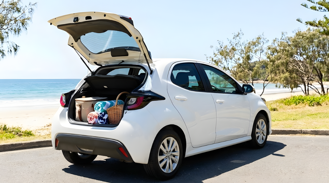 A cheerful, bright image of a modern, white compact hatchback (like a Hyundai i30 or Toyota Corolla) parked on a grassy patch near a beach or park. The boot (hatch) is open, revealing a simple picnic setup (cooler bag, towels). No people visible, just the suggestion of a day out