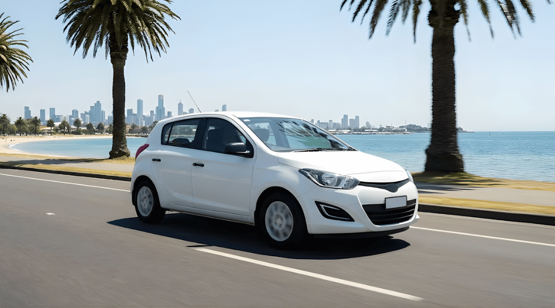 clean, white, unbranded compact hatchback driving along a scenic coastal road with palm trees (St Kilda/Beach Road vibe).