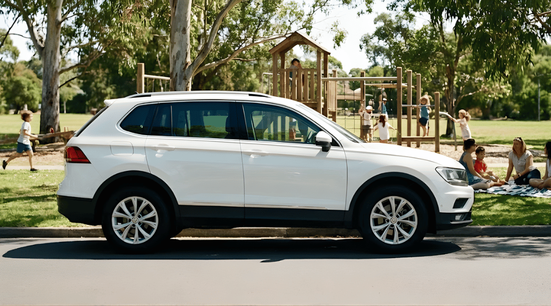 SUV parked near a grassy park or playground. In the background, children are playing actively (running or on play equipment) while parents relax on a picnic rug