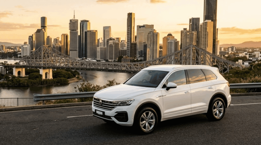 A pristine, clean, modern white passenger vehicle (sleek SUV) is parked smoothly in the foreground, angled to showcase its profile and the view. In the distance, the iconic structure of the Story Bridge is clearly visible, spanning the Brisbane River with the dense collection of modern glass CBD skyscrapers reflecting the warm, orange sunlight. The lighting is soft and golden, illuminating the car and the expansive city view.