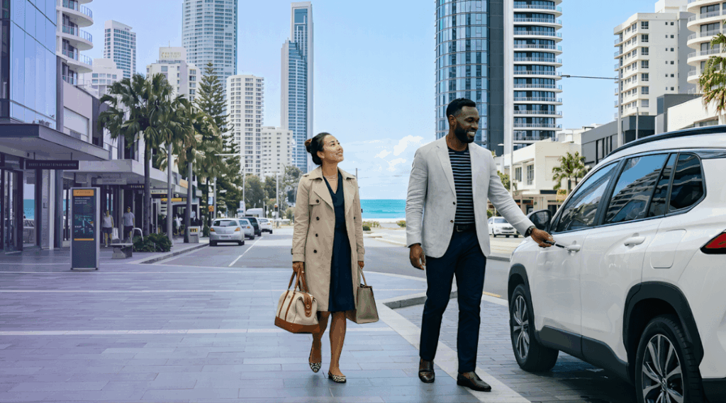 A sophisticated couple loading their sleek white rental SUV on a downtown Gold Coast street with modern skyscrapers and the beach strip in the background.